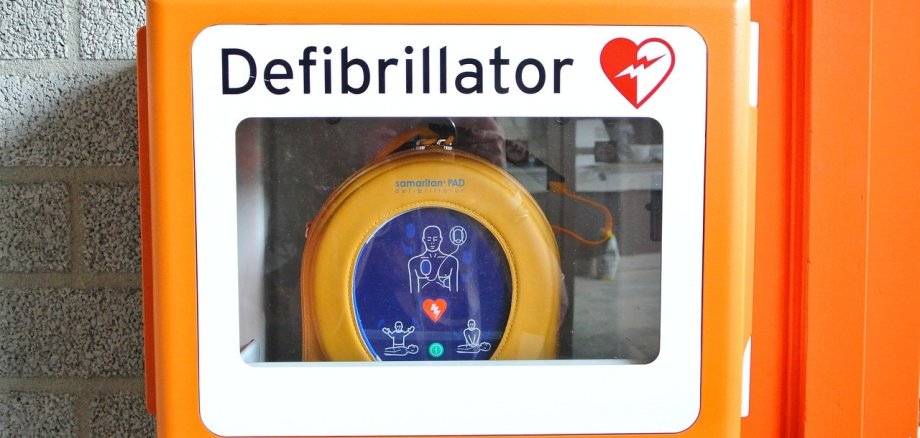 Auf dem Bild ist ein Defibrillator, der an einer gemauerten Wand hängt, abgebildet.